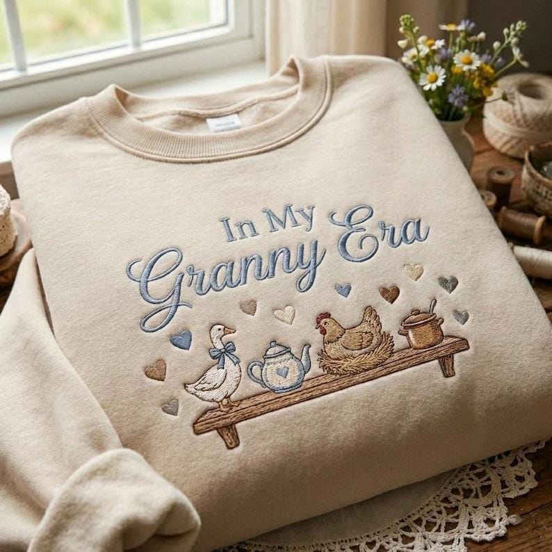 May include: Cream-coloured sweatshirt featuring the embroidered text "In My Granny Era" in blue. Below, an embroidered design shows a duck, teapot, chicken, and pot on a wooden shelf, with heart motifs.