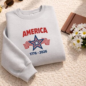 May include: A light gray sweatshirt with the word "AMERICA" in red letters above a blue and white star and red stripes. The text "1776-2026" is below the star. The sweatshirt is folded on a cream-colored surface.