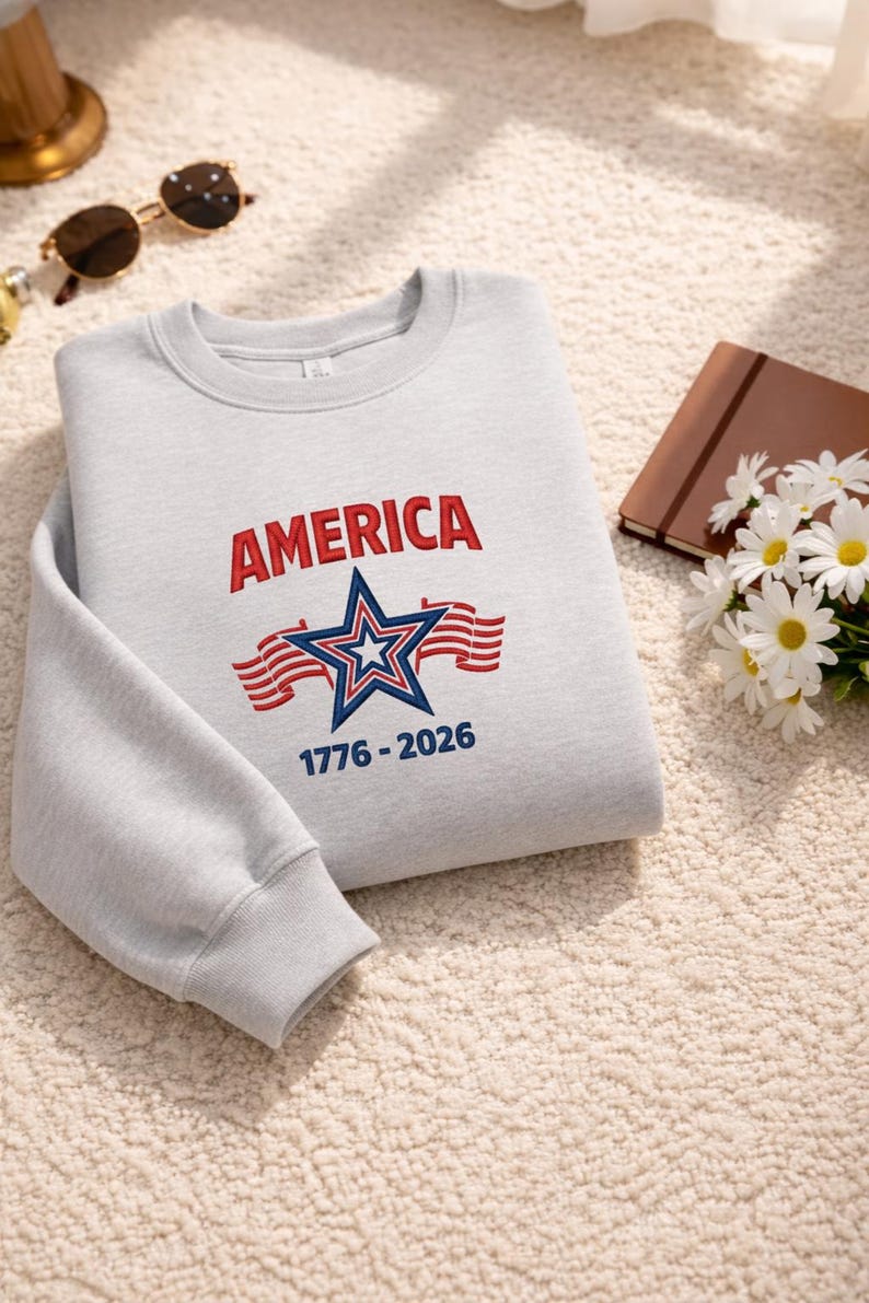 May include: A light gray sweatshirt with the word "AMERICA" in red letters above a blue and white star and red stripes. The text "1776-2026" is below the star. The sweatshirt is folded on a cream-colored surface.