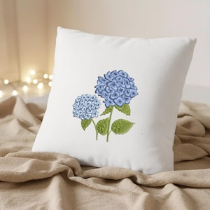 May include: A square white pillow featuring an embroidered design of two blue hydrangea flowers with green leaves and stems. The pillow is resting on a beige fabric surface, with soft lighting in the background.
