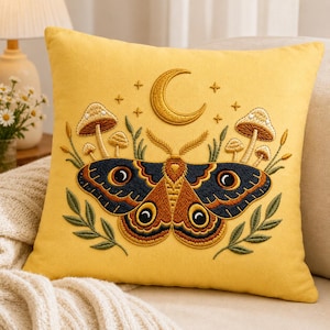 May include: Yellow decorative pillow with an embroidered design featuring a moth, mushrooms, a crescent moon, and stars. The moth has brown and navy blue wings with large eye-like markings. The pillow is square and sits on a beige surface.