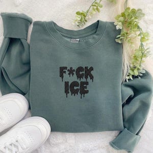 May include: A teal sweatshirt with the text "F*CK ICE" in a dripping black font. The sweatshirt has long sleeves and a crew neck. White sneakers are visible in the image.