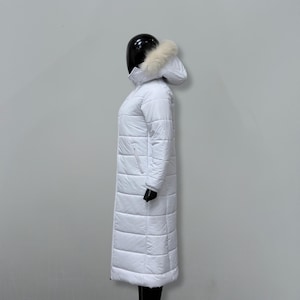 May include: A long, white puffer coat with a hood trimmed with faux fur. The coat is quilted and has a full-length design. The mannequin is wearing black gloves and pants.