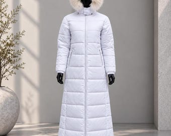 May include: A long, white, quilted puffer coat featuring a full-length front zipper, long sleeves, and a hood trimmed with a thick, fluffy, light beige faux fur. The coat has a straight, floor-length silhouette with horizontal stitching throughout.