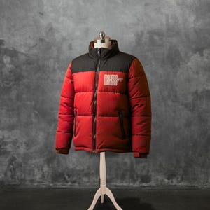 Puede incluir: Una chaqueta acolchada roja y negra con el texto "MAKERS PIT" en blanco. La chaqueta tiene un cuello alto, una cremallera de largo completo y dos bolsillos laterales. La chaqueta se exhibe en un maniquí.