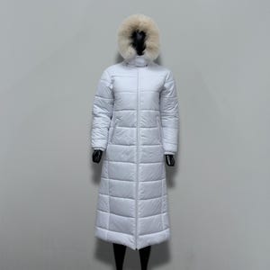 May include: A full-length, white puffer coat with a fur-trimmed hood. The coat features a vertical zipper closure and horizontal quilted stitching. The mannequin is wearing black gloves and boots. The coat is designed for cold weather.