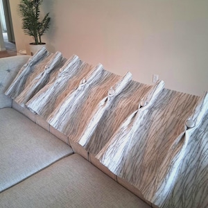 Peut inclure: Un canapé gris clair avec six coussins décoratifs. Les coussins présentent un motif à rayures blanches et grises et sont fixés avec de gros boutons ronds de couleur claire. Le canapé est de ton neutre et semble être fait d'un tissu texturé.