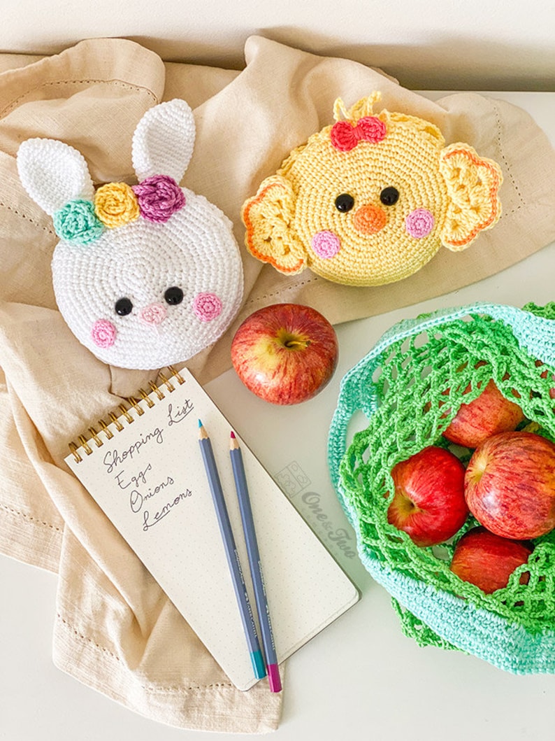 May include: Two round crochet pouches, one white rabbit with colorful flowers and one yellow chick with a red bow. A spiral-bound notepad with the handwritten text Shopping List, Eggs, Onions, Lemons, two colored pencils, a red apple, and a green net bag.