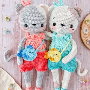 K&ouml;nnte beinhalten: Zwei geh&auml;kelte Katzenspielzeuge mit bunten Outfits und kleinen Taschen. Die Katze links tr&auml;gt ein rosa-rotes Outfit mit einer blauen Fisch-Tasche. Die Katze rechts tr&auml;gt ein blau-gr&uuml;nes Outfit mit einer gelben K&uuml;ken-Tasche.