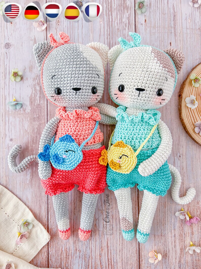 K&ouml;nnte beinhalten: Zwei geh&auml;kelte Katzenspielzeuge mit bunten Outfits und kleinen Taschen. Die Katze links tr&auml;gt ein rosa-rotes Outfit mit einer blauen Fisch-Tasche. Die Katze rechts tr&auml;gt ein blau-gr&uuml;nes Outfit mit einer gelben K&uuml;ken-Tasche.