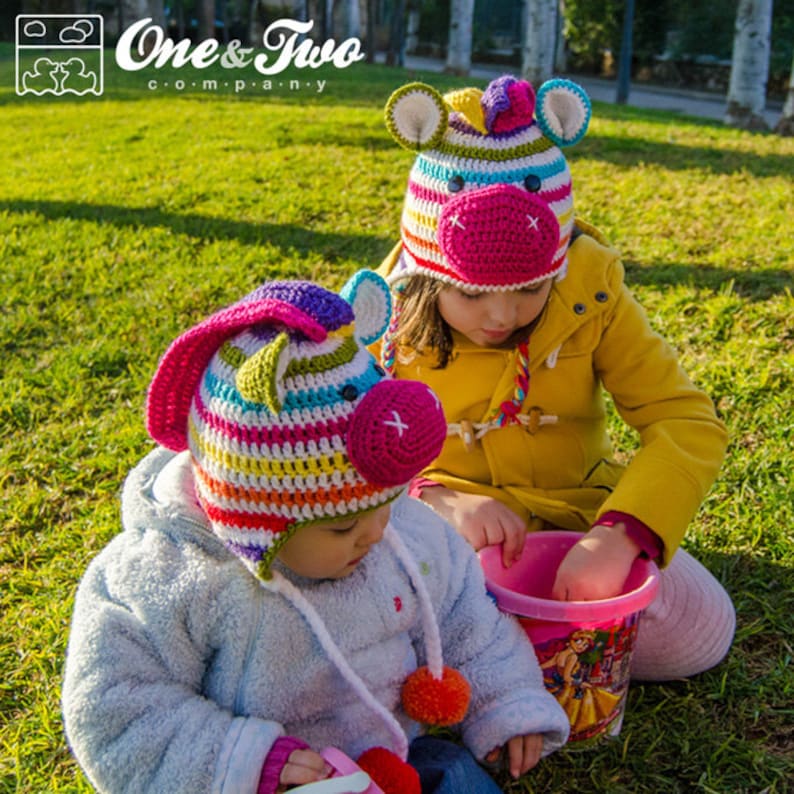 May include: Two children wearing colorful, handmade crochet hats shaped like unicorns. The hats feature striped patterns in various colors, including pink, blue, green, and yellow. The hats have large ears and a pink snout. The image includes the "One & Two Company" logo.