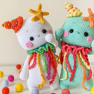 May include: Two crocheted amigurumi toys with colorful yarn accents. One is white with a red, orange, yellow, and green scarf and a starfish on its head. The other is teal with a pink, yellow, and orange scarf and a starfish on its head.