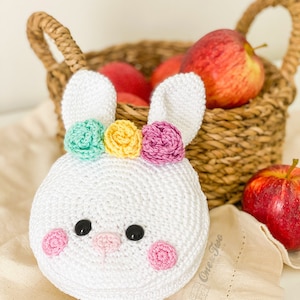 May include: A white crocheted bunny head with two long ears, featuring three small flowers in mint green, yellow, and pink on top. The face has two black circular eyes, a small pink nose, and two pink circular cheeks. The text One Two is visible.