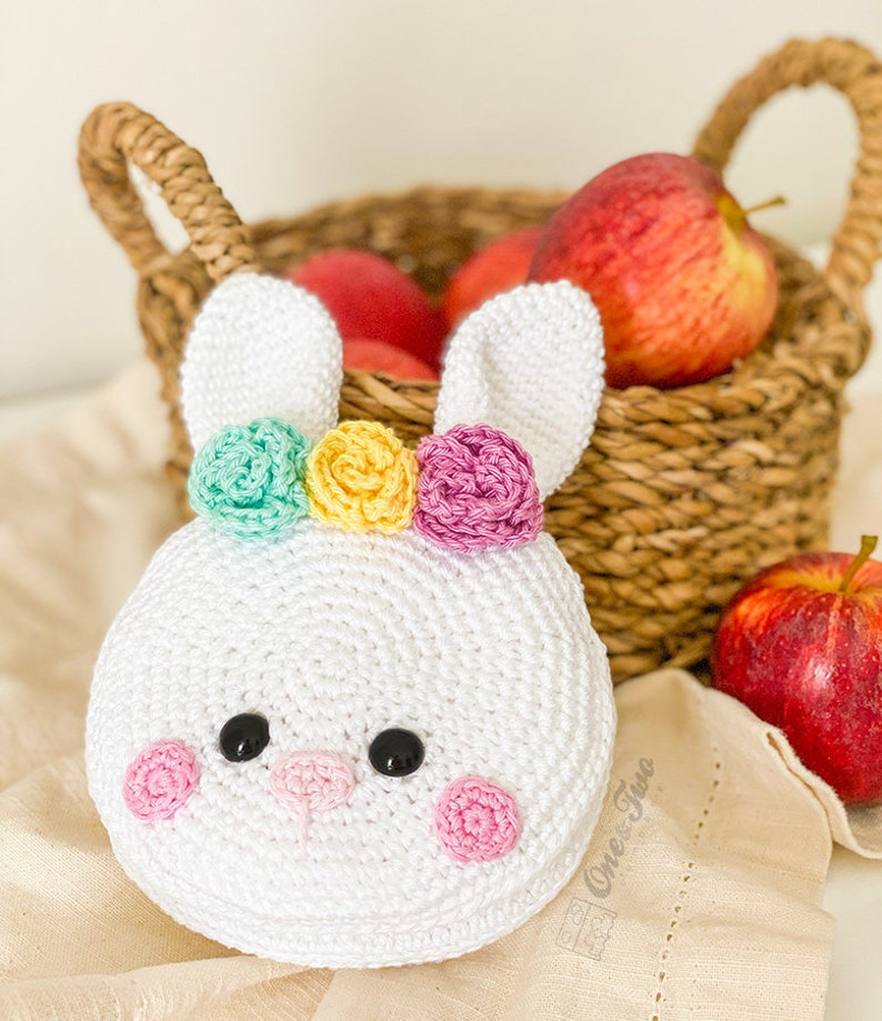 May include: A white crocheted bunny head with two long ears, featuring three small flowers in mint green, yellow, and pink on top. The face has two black circular eyes, a small pink nose, and two pink circular cheeks. The text One Two is visible.