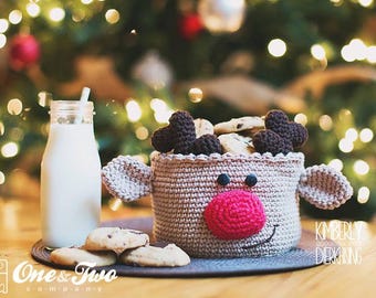 May include: A crocheted reindeer-shaped basket filled with chocolate chip cookies. The basket is beige with a pink nose and brown antlers. A glass bottle of milk sits on a wooden table next to the basket.