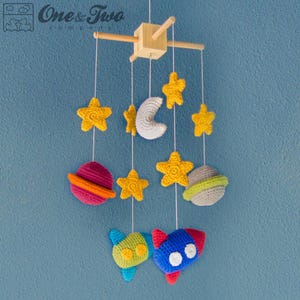 May include: A wooden baby mobile with crocheted planets, stars, and a rocket. The planets are red, green, and gray. The stars are yellow. The rocket is blue and green.