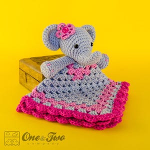 May include: A crocheted elephant toy with a pink flower on its head, holding a pink and gray crocheted baby blanket. The blanket has a scalloped edge.