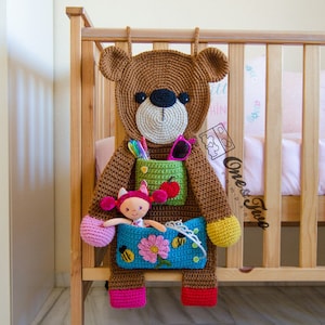 Puede incluir: Un oso de peluche de crochet marrón con un bolsillo verde, rojo y amarillo en el pecho. El bolsillo tiene una manzana roja y una pequeña muñeca dentro. El oso está colgado en una barandilla de cuna de madera.