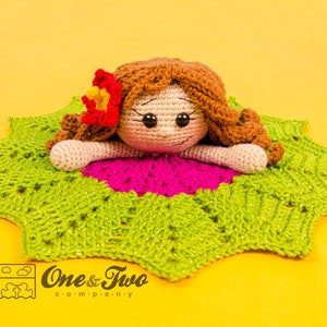 Puede incluir: Una mu&ntilde;eca de crochet con cabello casta&ntilde;o y una flor rosa en la cabeza est&aacute; acostada sobre una manta de crochet verde y rosa. La manta tiene forma de estrella. La mu&ntilde;eca tiene una expresi&oacute;n dulce en su rostro. El texto "One & Two Company" es visible en la esquina inferior derecha de la imagen.