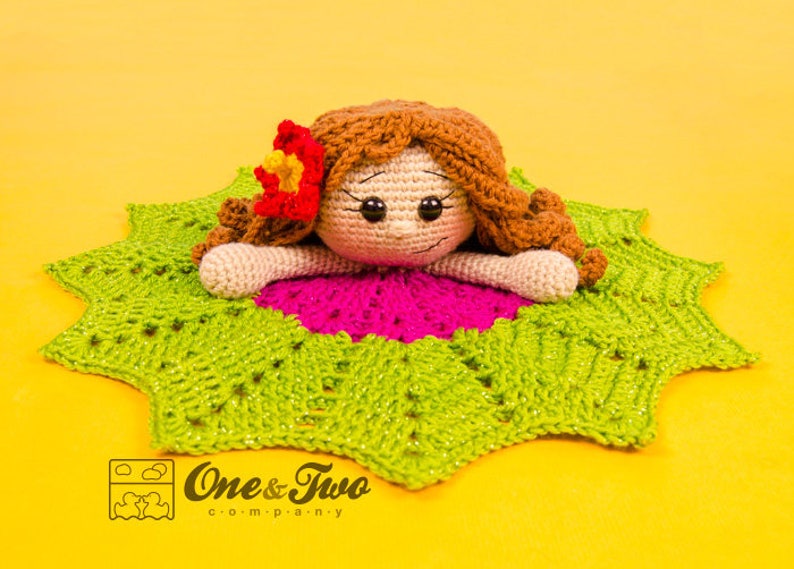 Puede incluir: Una mu&ntilde;eca de crochet con cabello casta&ntilde;o y una flor rosa en la cabeza est&aacute; acostada sobre una manta de crochet verde y rosa. La manta tiene forma de estrella. La mu&ntilde;eca tiene una expresi&oacute;n dulce en su rostro. El texto "One & Two Company" es visible en la esquina inferior derecha de la imagen.