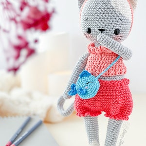 K&ouml;nnte beinhalten: Eine geh&auml;kelte graue und wei&szlig;e Katzendolle, die ein rosa Kleid und eine blaue Handtasche tr&auml;gt. Die Puppe hat eine rosa Schleife auf dem Kopf und schwarze Augen. Die Puppe steht auf einer wei&szlig;en Oberfl&auml;che.