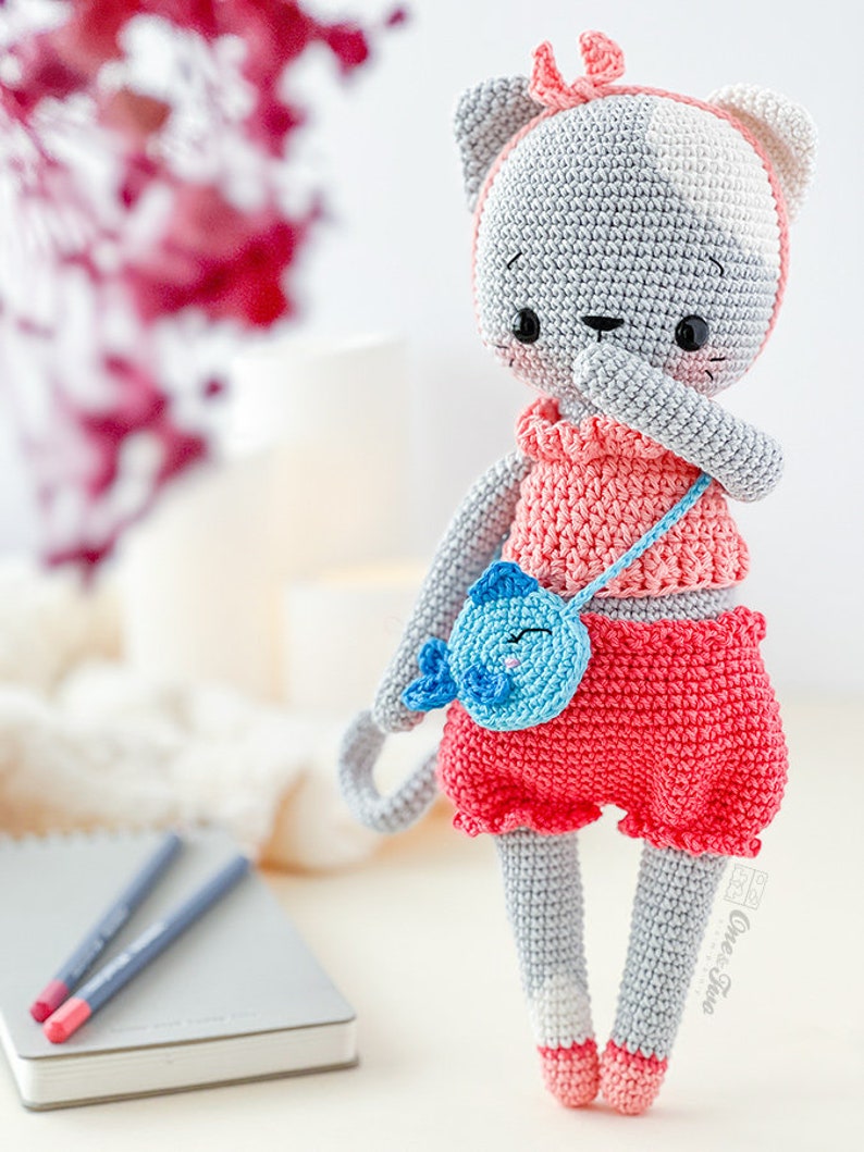 K&ouml;nnte beinhalten: Eine geh&auml;kelte graue und wei&szlig;e Katzendolle, die ein rosa Kleid und eine blaue Handtasche tr&auml;gt. Die Puppe hat eine rosa Schleife auf dem Kopf und schwarze Augen. Die Puppe steht auf einer wei&szlig;en Oberfl&auml;che.