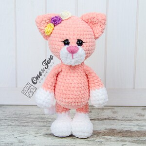 May include: A pink crocheted cat with white feet and a white nose. The cat has yellow, purple, and white flowers on its head. The cat is standing on a wooden surface.