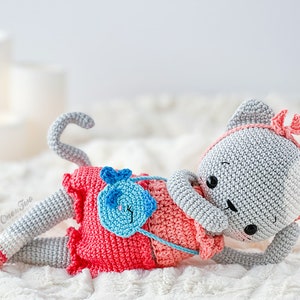 K&ouml;nnte beinhalten: Ein geh&auml;keltes graues Katzenspielzeug mit rosa und blauen Akzenten. Die Katze tr&auml;gt ein rosa Kleid und eine blaue Handtasche. Die Katze liegt auf einer wei&szlig;en, flauschigen Oberfl&auml;che.
