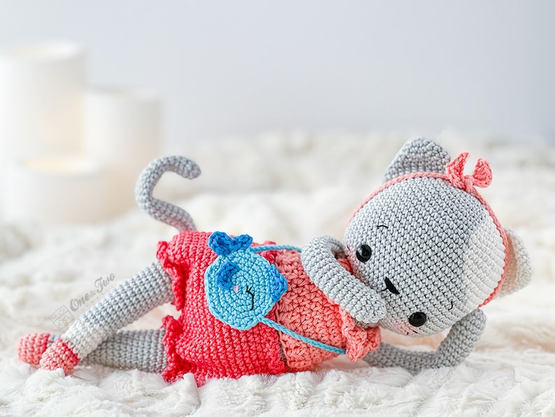 K&ouml;nnte beinhalten: Ein geh&auml;keltes graues Katzenspielzeug mit rosa und blauen Akzenten. Die Katze tr&auml;gt ein rosa Kleid und eine blaue Handtasche. Die Katze liegt auf einer wei&szlig;en, flauschigen Oberfl&auml;che.