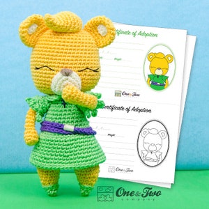 May include: A crocheted stuffed animal bear wearing a green dress with a purple belt. The bear has yellow fur, a white nose, and closed eyes. The bear is standing on a light blue background. The image also includes a certificate of adoption with the text "One & Two Company" printed on it.