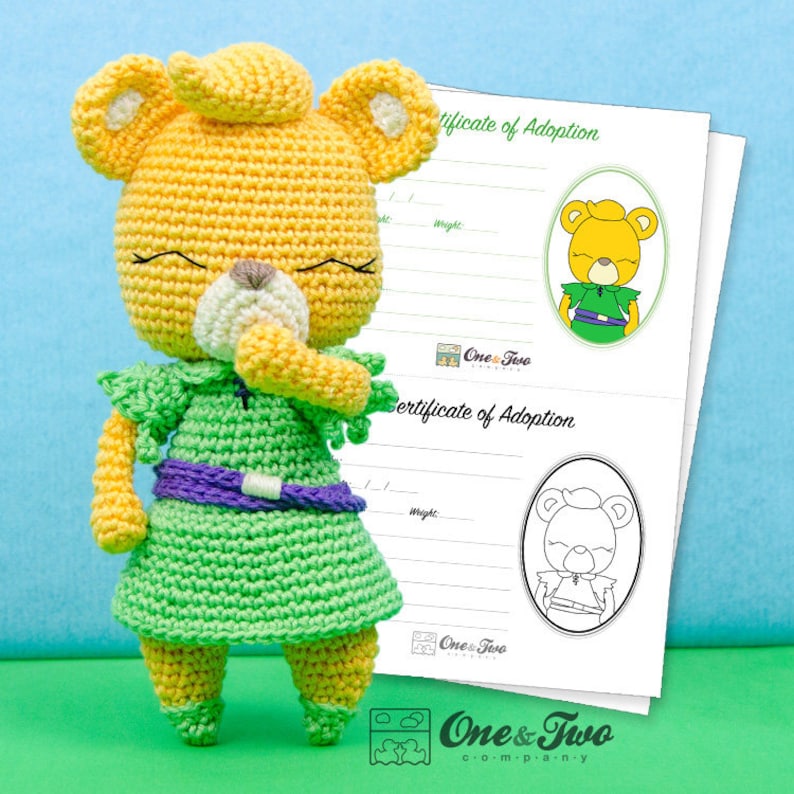 May include: A crocheted stuffed animal bear wearing a green dress with a purple belt. The bear has yellow fur, a white nose, and closed eyes. The bear is standing on a light blue background. The image also includes a certificate of adoption with the text "One & Two Company" printed on it.