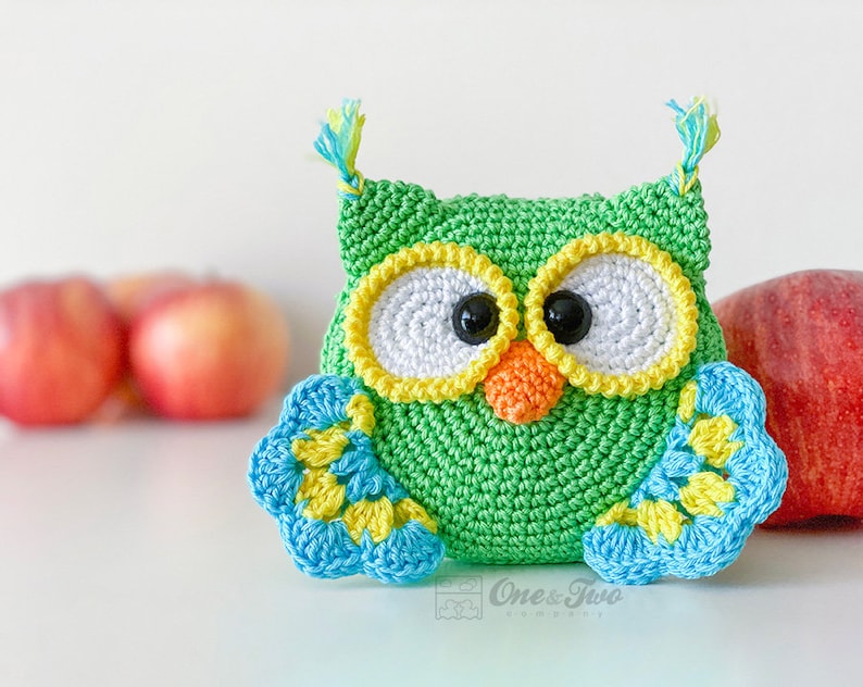 May include: A green crocheted owl with large white eyes, yellow borders, and black pupils. It features an orange beak, blue and yellow wings, and small tufts on top. The text One & Two Company is printed in a small box at the bottom centre.