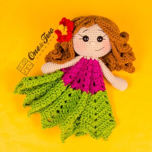 Puede incluir: Una mu&ntilde;eca de crochet con cabello casta&ntilde;o, un vestido rosa y verde y una flor en el pelo. La mu&ntilde;eca est&aacute; tumbada sobre un fondo amarillo.
