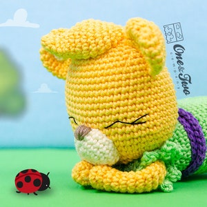 May include: A yellow crocheted stuffed animal with green and purple accents, lying on its side with its eyes closed. A red ladybug is in the foreground.