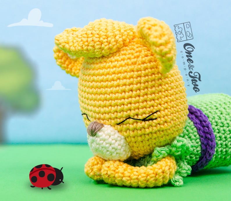 May include: A yellow crocheted stuffed animal with green and purple accents, lying on its side with its eyes closed. A red ladybug is in the foreground.