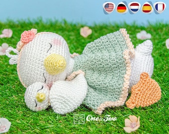 Könnte beinhalten: Eine weiße Häkel-Amigurumi-Puppe mit einem grünen Kleid und einer rosa Blumenkrone. Die Puppe hält eine kleinere weiße Häkel-Puppe mit einem gelben Schnabel. Die Puppen liegen auf einem Bett aus grünem Gras.