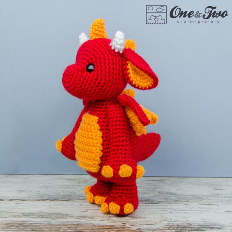 K&ouml;nnte beinhalten: Ein roter H&auml;kel-Drache mit gelben Akzenten und wei&szlig;en H&ouml;rnern. Der Drache steht auf einer Holzoberfl&auml;che.