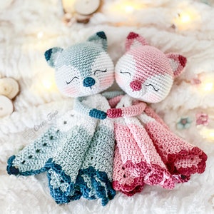 May include: Two crocheted fox dolls, one in shades of blue and the other in pink, are nestled together. The dolls have closed eyes, rosy cheeks, and detailed features. Their blankets have scalloped edges, adding a delicate touch to the handmade toys.