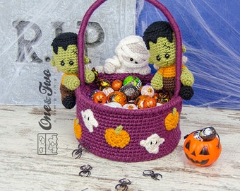 May include: A purple crocheted Halloween trick-or-treat basket with a handle, filled with candy and decorated with ghosts and pumpkins. Two crocheted figures, a mummy and a Frankenstein, are holding onto the basket. There are also small plastic spiders on the table.