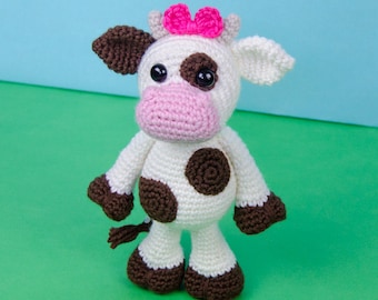 Puede incluir: Una vaca de crochet blanca con manchas marrones y un lazo rosa. La vaca tiene ojos negros y una nariz rosa. La vaca está de pie sobre una superficie verde.