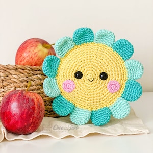 May include: A round, crocheted flower with a yellow center, black bead eyes, a small stitched smile, and pink cheeks. The petals alternate between light teal and dark teal. The text One and Two is visible at the bottom of the image.