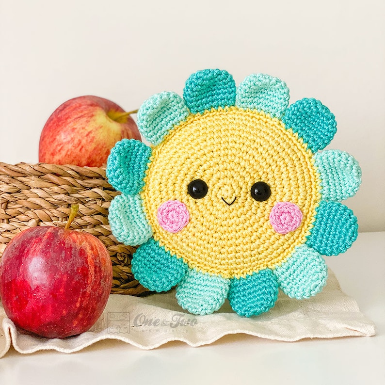 May include: A round, crocheted flower with a yellow center, black bead eyes, a small stitched smile, and pink cheeks. The petals alternate between light teal and dark teal. The text One and Two is visible at the bottom of the image.