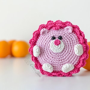 May include: A round, crocheted animal figure in light pink with a dark pink ruffled border. It features a white muzzle, a dark pink nose, two black bead eyes, and small white paws. The text "Ones & Two" is printed in a light grey font on the lower left.