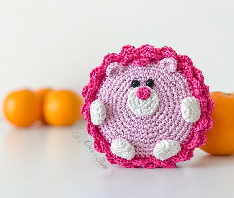 May include: A round, crocheted animal figure in light pink with a dark pink ruffled border. It features a white muzzle, a dark pink nose, two black bead eyes, and small white paws. The text "Ones & Two" is printed in a light grey font on the lower left.