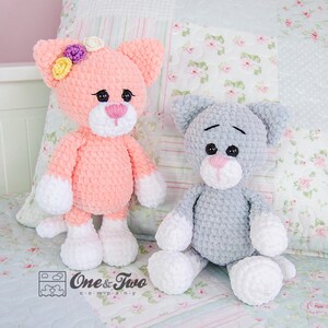 May include: Two crocheted cat toys, one pink with white accents and a flower crown, and one grey with white accents. Both are sitting on a floral patterned fabric.