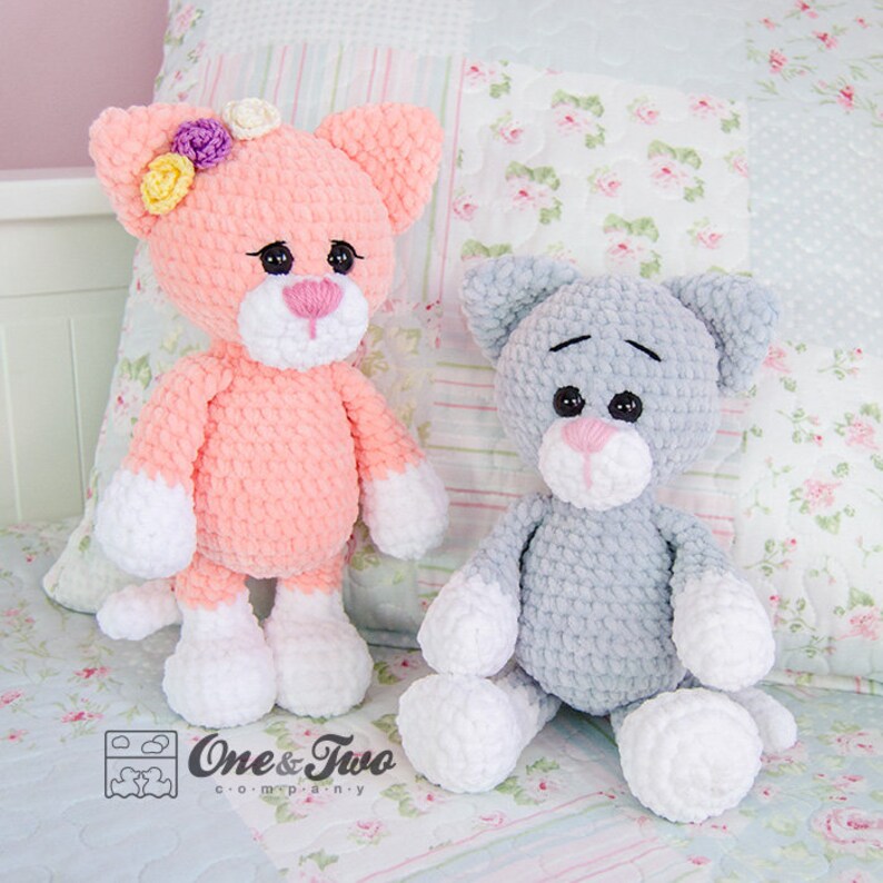 May include: Two crocheted cat toys, one pink with white accents and a flower crown, and one grey with white accents. Both are sitting on a floral patterned fabric.