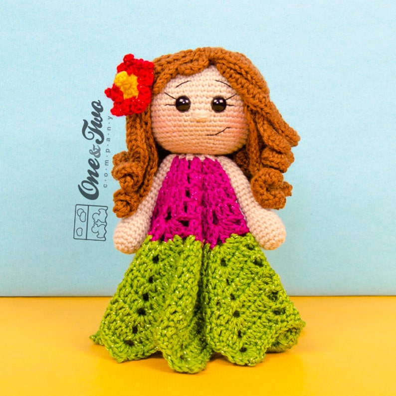 Puede incluir: Una mu&ntilde;eca de crochet con pelo marr&oacute;n, un vestido rosa y verde y una flor roja en la cabeza.
