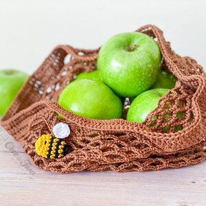 May include: A brown crocheted mesh market bag containing several bright green apples. A small, crocheted bee applique with yellow, black, and white yarn is attached to the front of the bag. The text One & Two is visible on the lower left side.