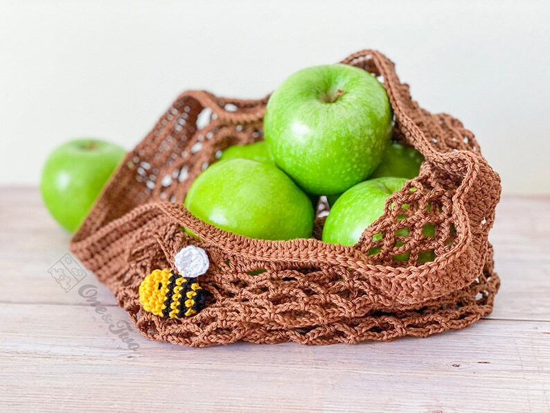 May include: A brown crocheted mesh market bag containing several bright green apples. A small, crocheted bee applique with yellow, black, and white yarn is attached to the front of the bag. The text One & Two is visible on the lower left side.
