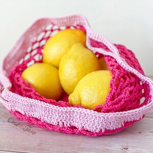 May include: A bright pink and light pink crocheted net bag containing four yellow lemons. The bag features a wide, solid crocheted border at the top and a mesh body. A small logo with the text One & Two is visible on the lower left side.